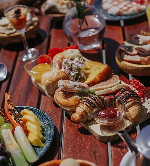 Brunch servido em uma mesa de madeira. Inclui frutas, doces e carnes, com um vaso de vidro com flores.