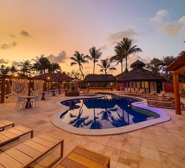 Piscina ao pôr do sol com palmeiras, espreguiçadeiras e áreas de estar cobertas.