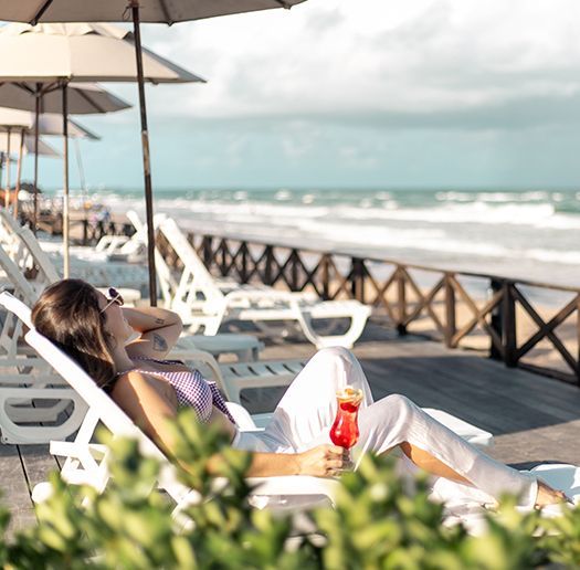 Mulher relaxando em uma cadeira de praia, segurando uma bebida, com o oceano ao fundo.