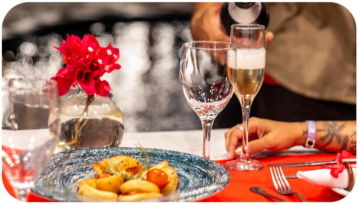 Garçom servindo champanhe em uma mesa de jantar posta para dois, toalha de mesa vermelha, piscina e luzes ao fundo.