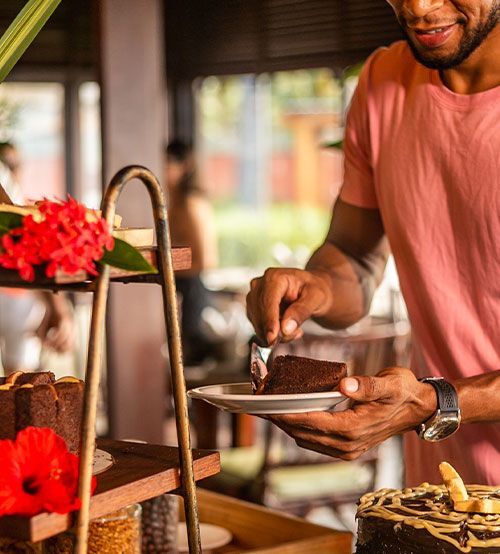 Homem pegando uma fatia de bolo de chocolate de um bufê. Ao ar livre, luz do sol, camisa rosa.