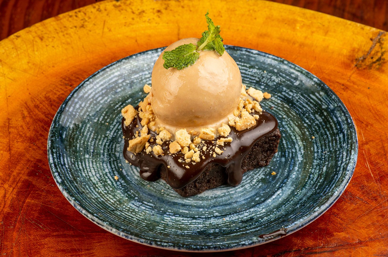 Close de sobremesa em um prato azul, com base de biscoito esfarelado, decorada com folhas de hortelã. Uma mão segurando uma colher.