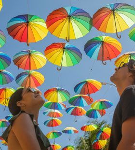 Pessoas olhando para um céu repleto de guarda-chuvas coloridos com listras do arco-íris. Ao fundo, céu azul.