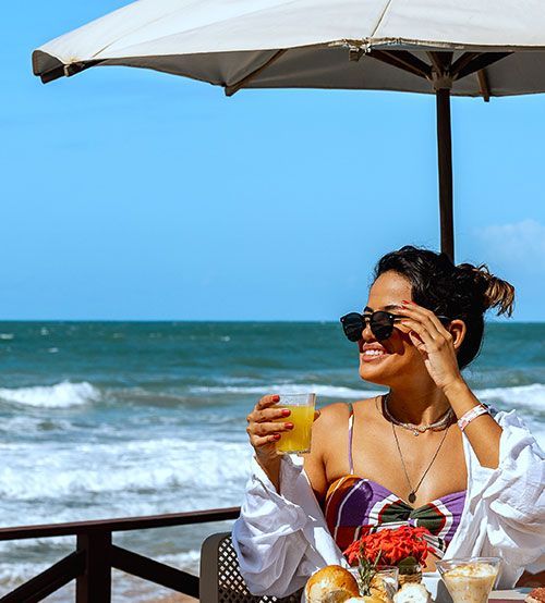 Mulher sorrindo em uma mesa na praia, segurando uma bebida, sob um guarda-chuva com o oceano ao fundo.