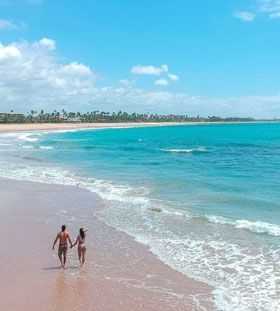 Casal caminhando na praia sob o sol; água e céu azuis; costa arenosa.