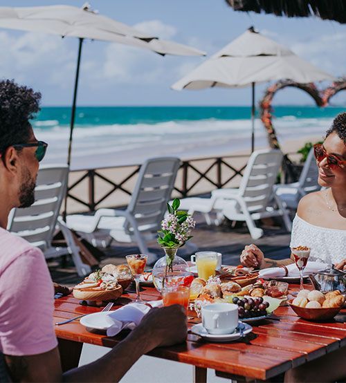 Casal desfrutando de um café da manhã na praia com frutas, pães e bebidas em uma mesa de madeira.