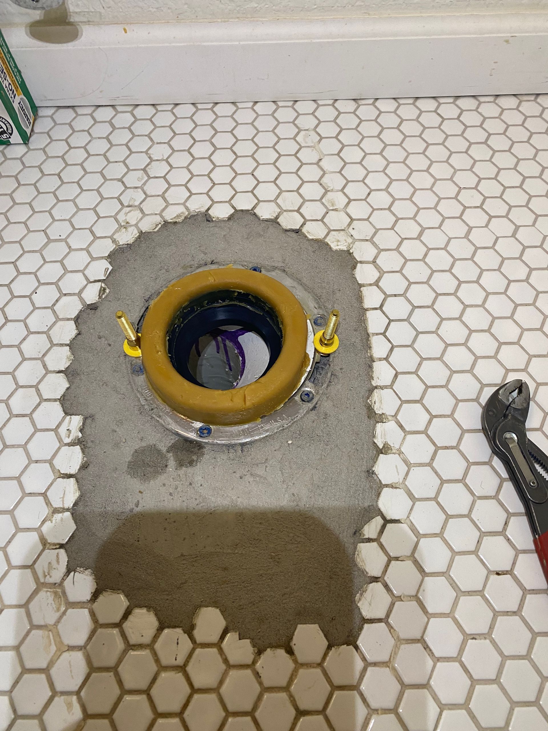 A toilet flange is sitting on a tiled floor next to a pair of pliers.