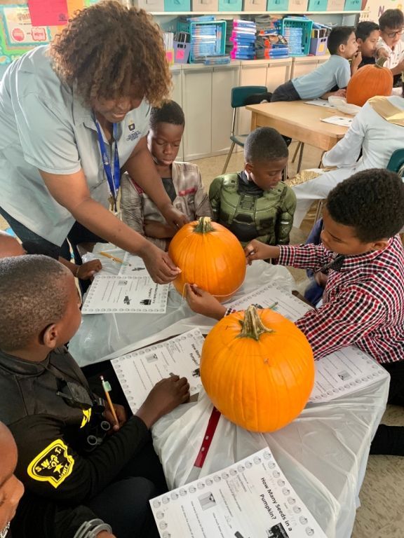 A woman is teaching a group of children how to carve pumpkins