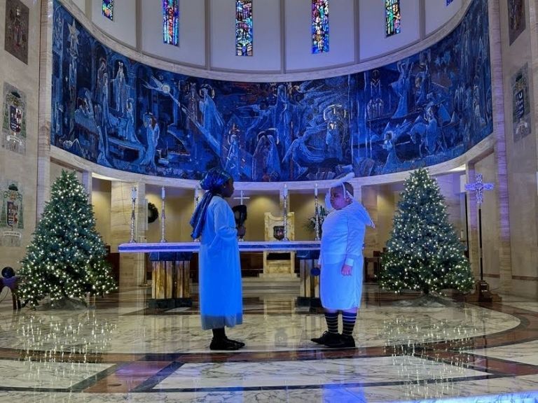 Two women in blue coats are standing in a church with christmas trees.