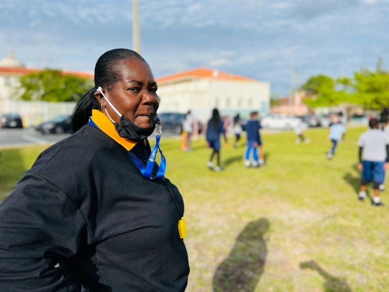 A woman wearing a mask is standing in a field with children playing in the background.
