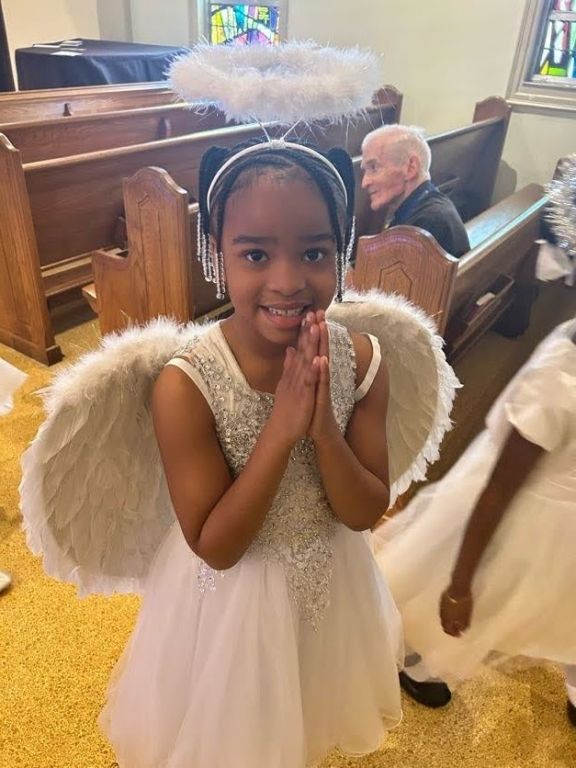 A little girl in a white dress and angel wings is praying in a church.