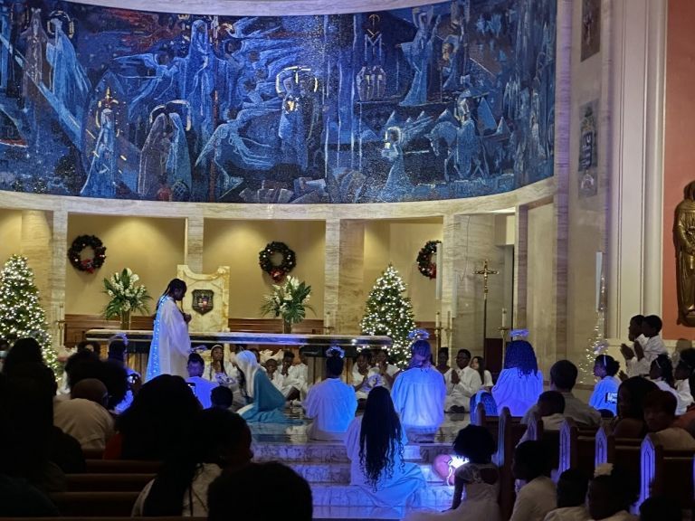 A group of people are sitting in a church with christmas trees in the background