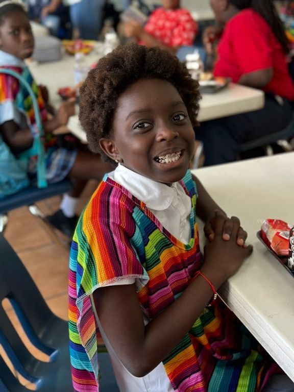 A young girl in a colorful dress is smiling while sitting at a table.