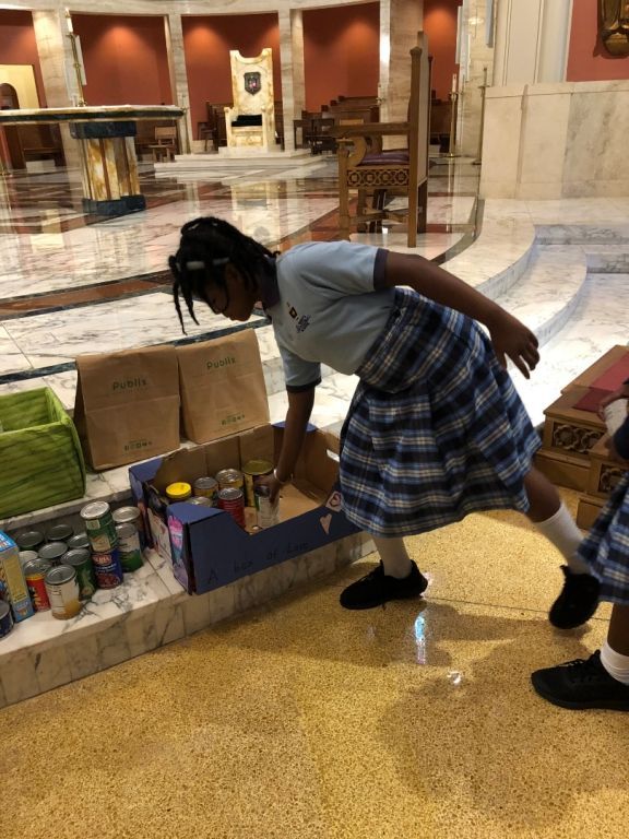 A girl in a plaid skirt is reaching into a box of food in a church.