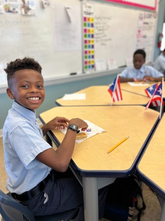A young boy is smiling while sitting at a desk in a classroom