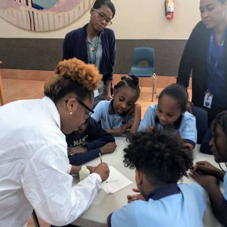 A group of children are sitting around a table with a woman writing on a piece of paper