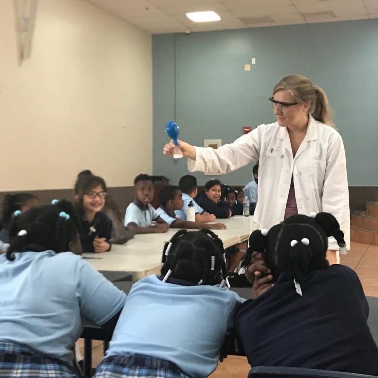 A woman in a lab coat is holding a blue object in front of a group of children
