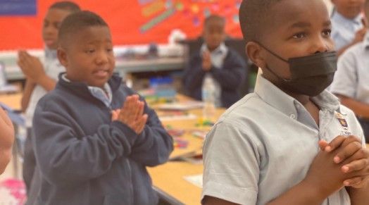 A group of young boys wearing masks are clapping their hands in a classroom.