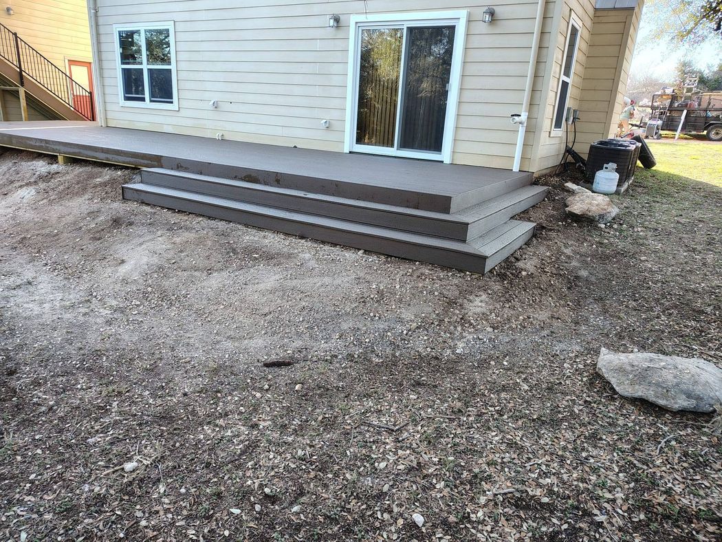 A gray deck with three wide steps leads to a beige house with a sliding glass door and a window, set in a dirt yard.