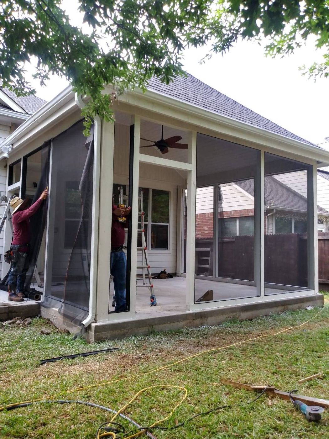 Two workers install large window screens into a cream-colored outdoor gazebo frame on a grassy lawn.