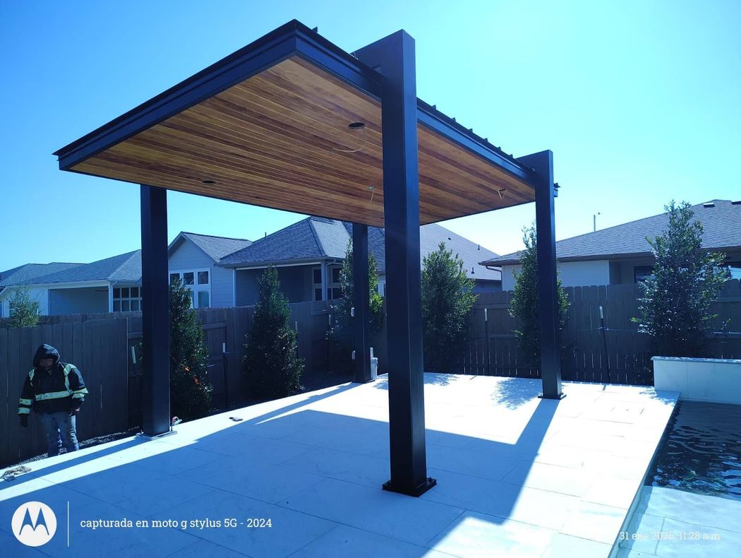 A modern black-framed patio pavilion with a wooden ceiling stands over a light-colored paved area in a backyard.