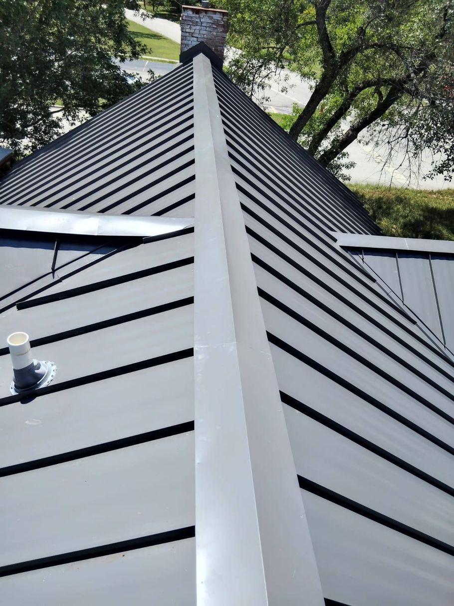 A top-down view of a dark metal roof ridge, showing the seam and surrounding panels leading to a brick chimney.