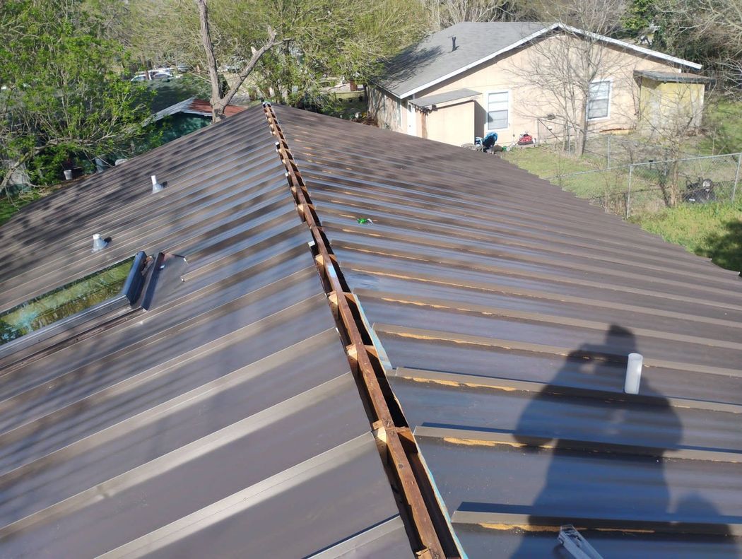 A high-angle view of a brown metal roof under repair, showing an open ridge vent strip and the shadow of the photographer.
