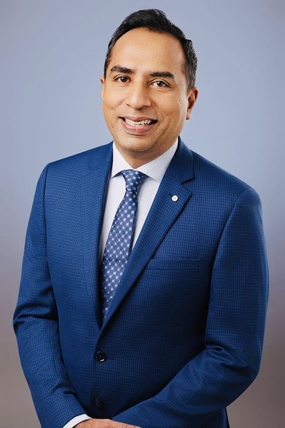 Man in a blue suit smiling, wearing a patterned tie, standing against a light blue backdrop.