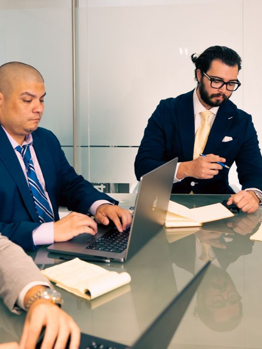 Three men in suits at a conference table with laptops and notepads, discussing work.