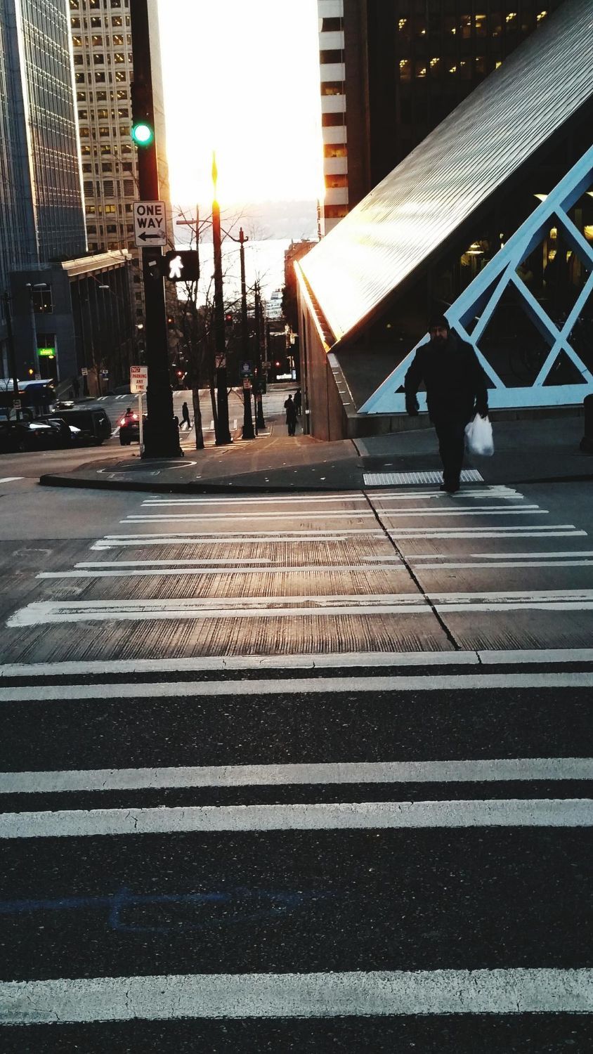 Person crossing a crosswalk in a city. Buildings and setting sun are in the background.