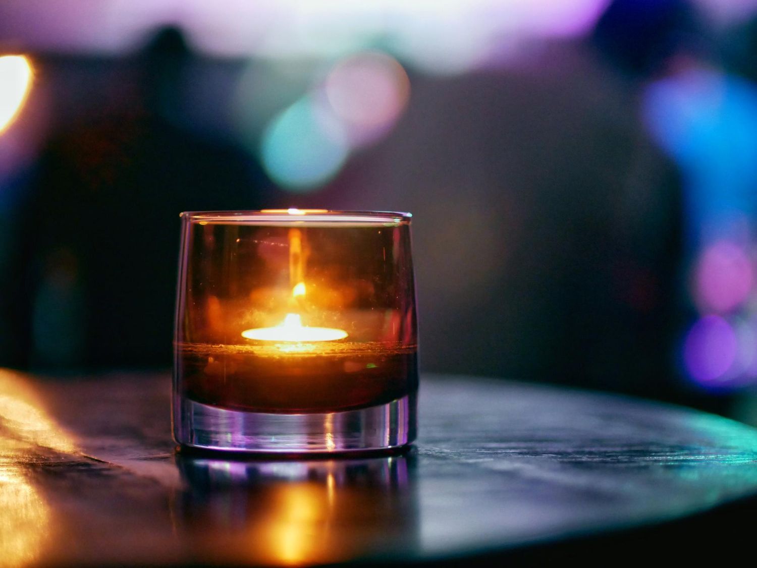 Candle flame in a glass holder on a dark, reflective surface, with blurry colorful lights in the background.