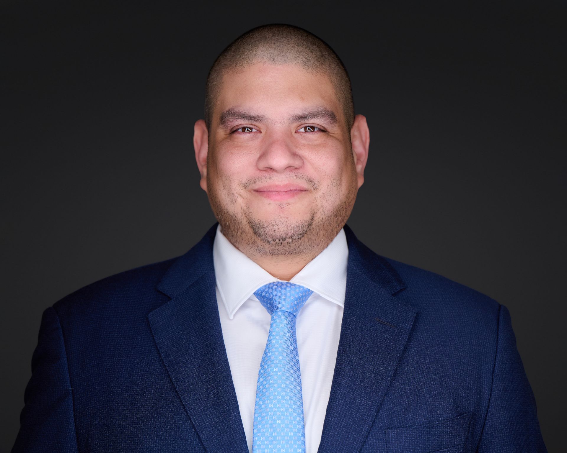 Man in a suit smiles at the camera, seated at a desk with a laptop.