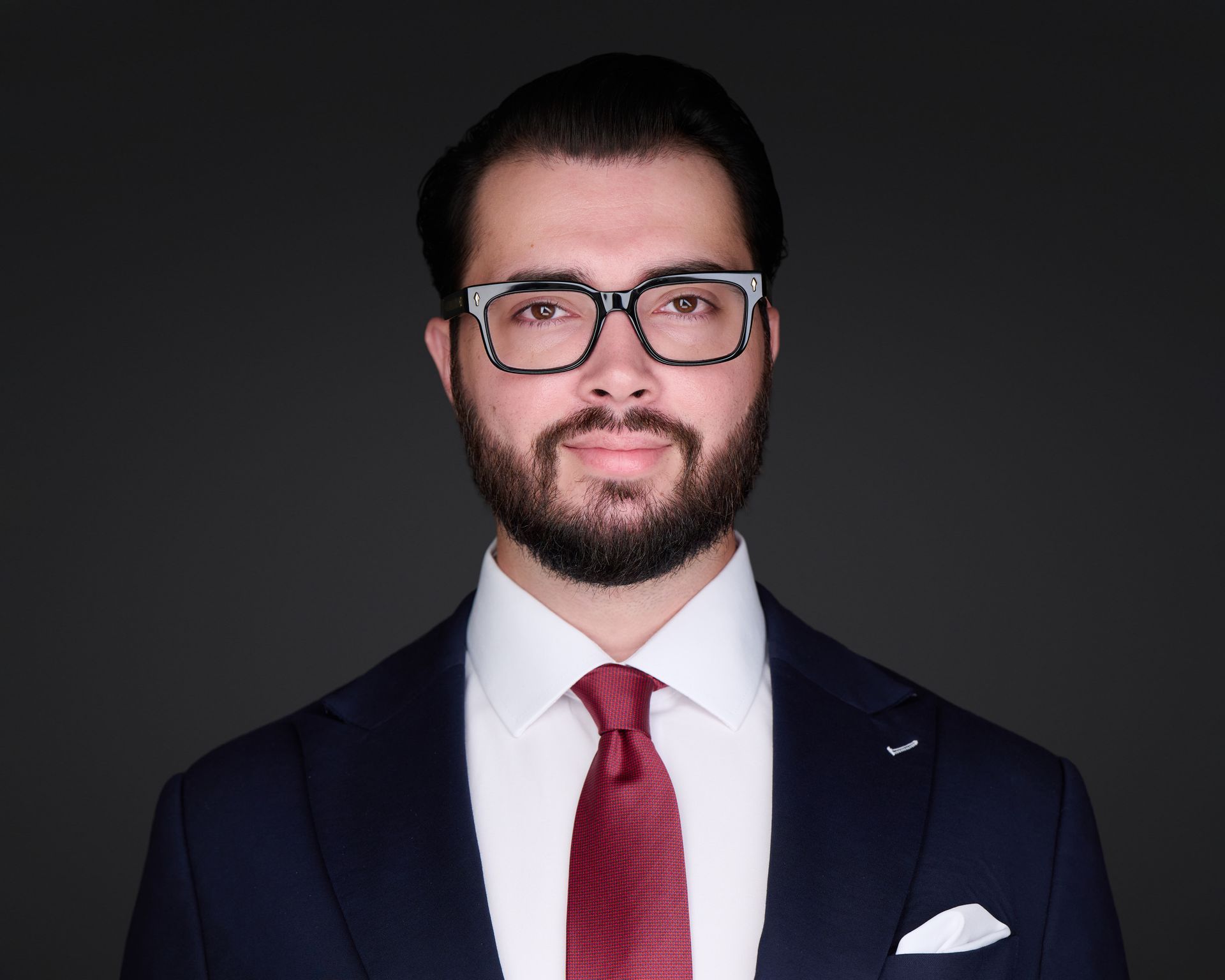 Man in navy suit, glasses, and red tie, smiling. Dark gray background.
