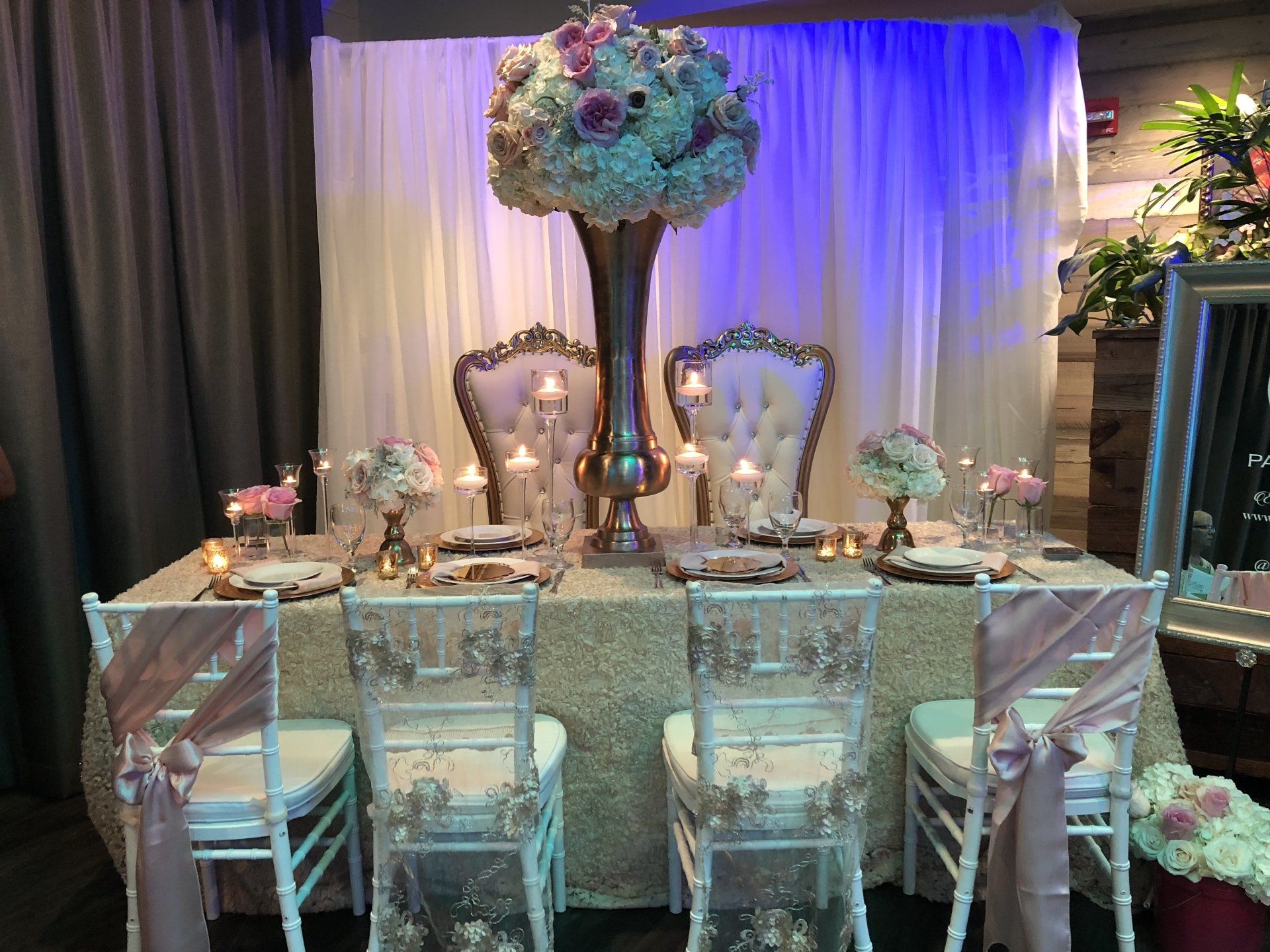 Table set up with floral centerpieces (two small and one tall), two thrones on one side and event chairs on the other side with gorgeous covers.