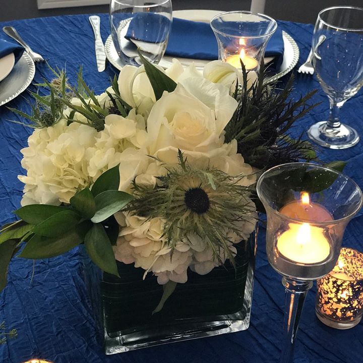 Floral arrangement centerpiece in a short square class vase sitting on a crushed blue table cloth.