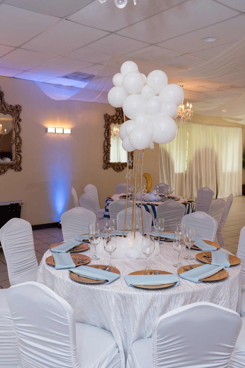 An elegant contemporary centerpiece with a gold four post curved frame, white balloons of various sizes at the top, hanging stars and a cotton cloud at the base.
