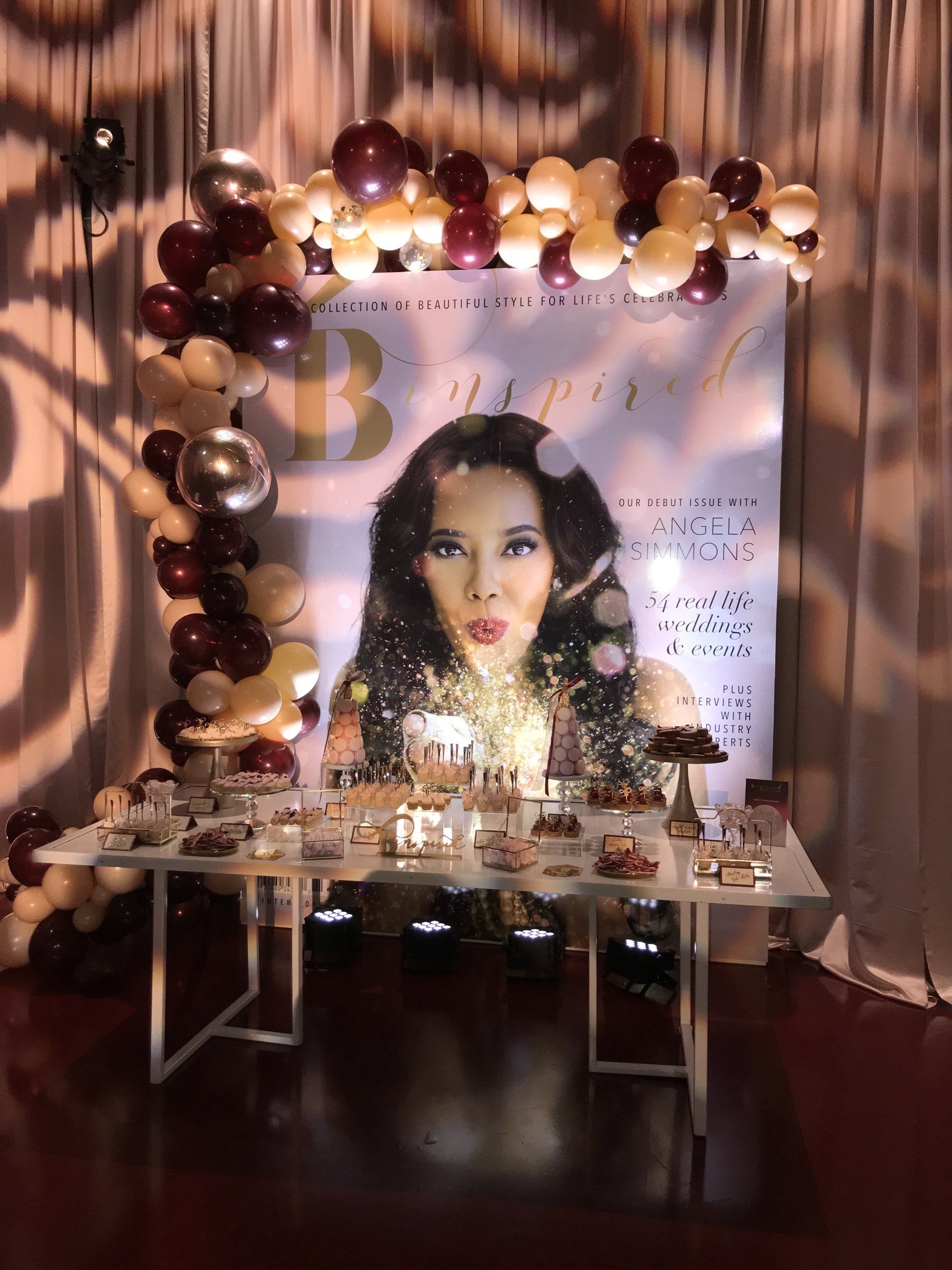 A classy display table with a magazine cover style backdrop framed by an organic balloon garland.