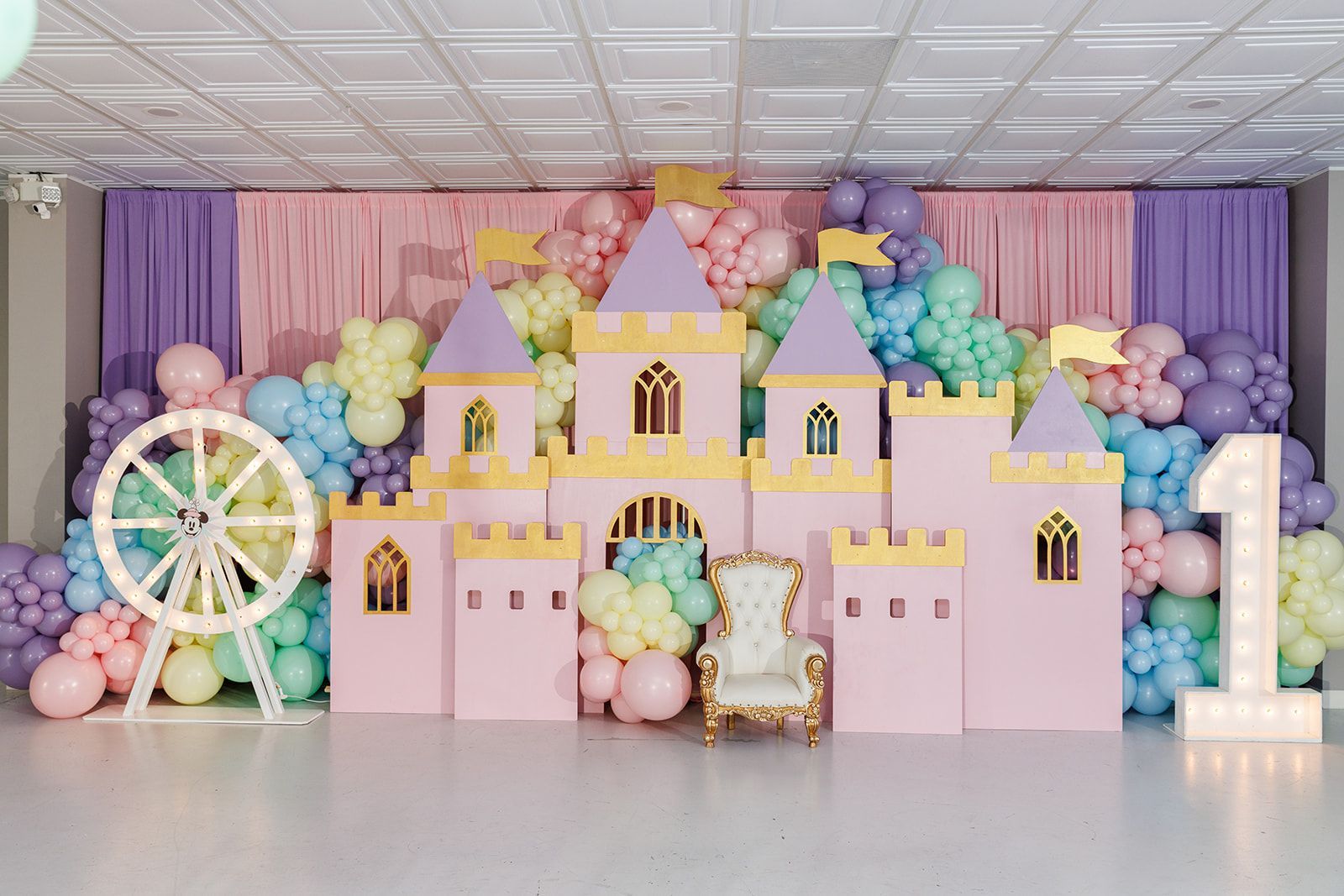Pastel princess castle backdrop with rainbow balloon garlands and light-up number one for a kids birthday party setup