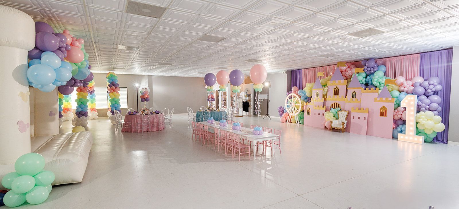 Wide view of pastel kids party venue with long tables, balloon garlands, and princess castle backdrop installation