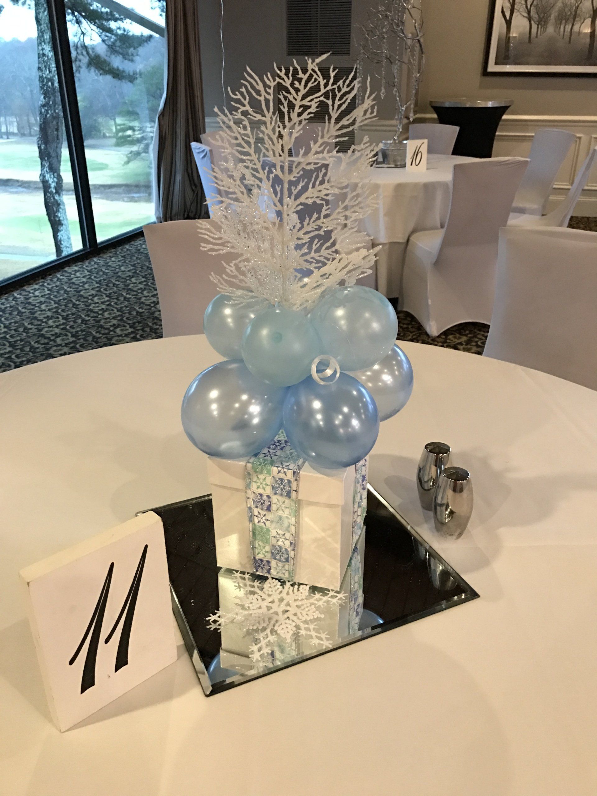 Winter themed centerpieces with a present box at the bottom blue balloons and a sparkly white tree at the top.