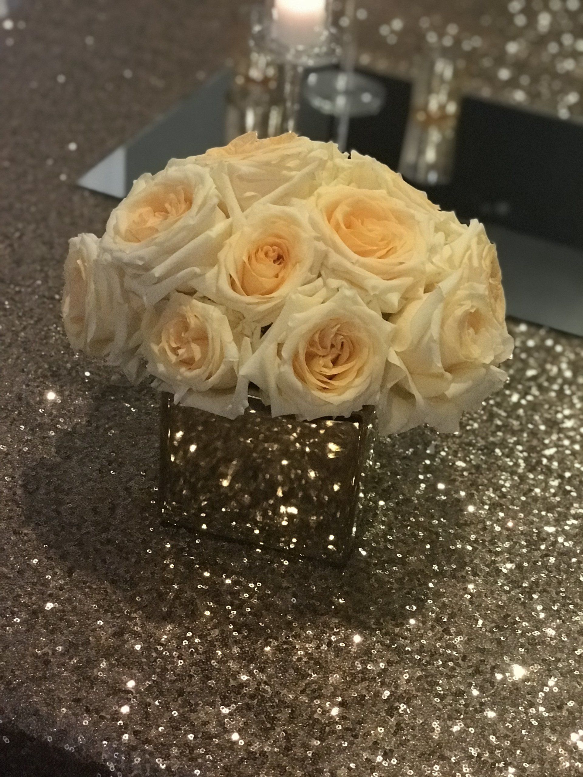 A centerpiece created with white roses in a gold cube vase on a gold sequin table cloth.