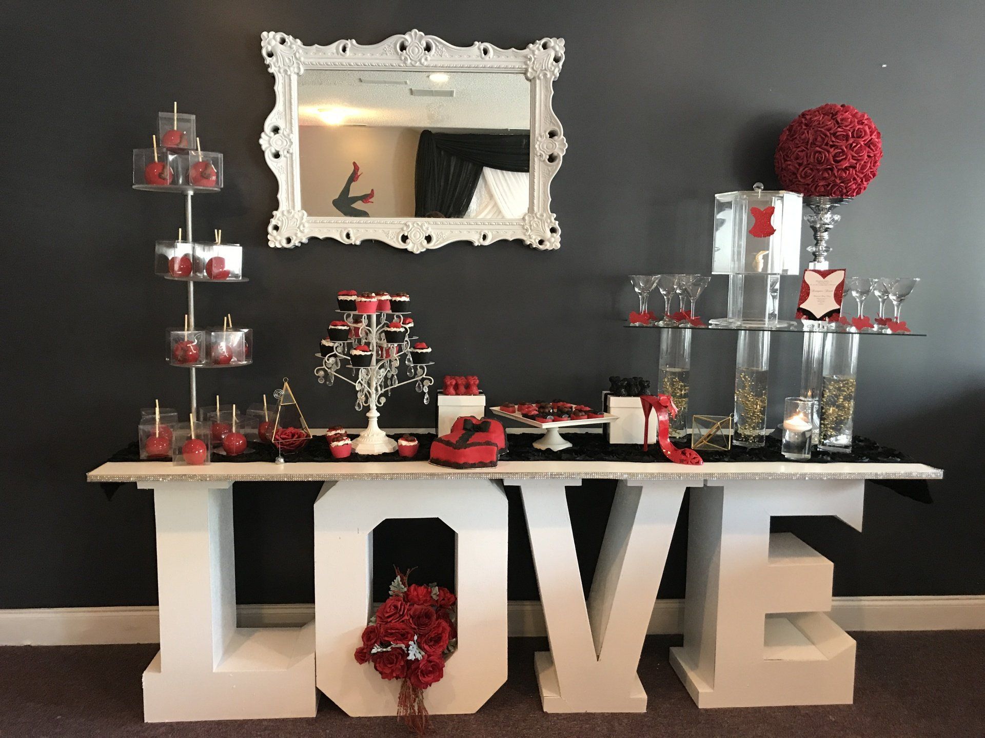 Dessert table with a red, black and white theme, elegant props and a white mirror on the wall behind. Table top is sitting on large 