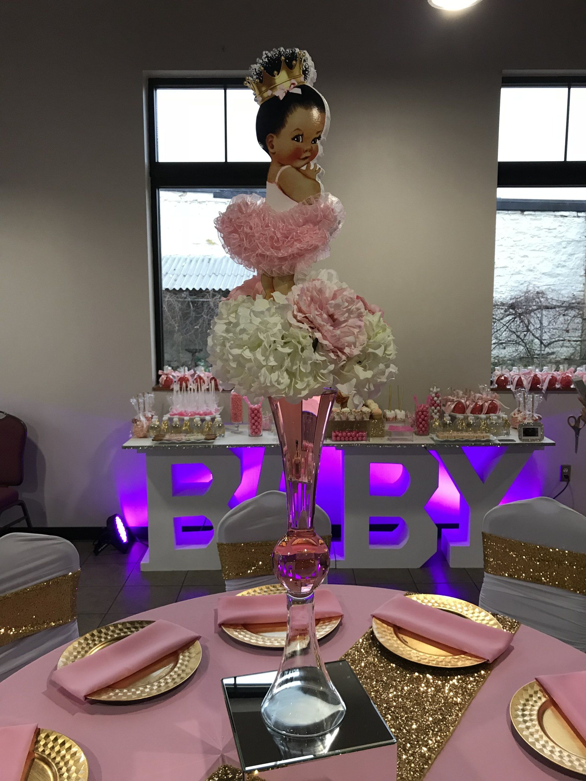 Custom baby shower event design with elegant chair covers, pink table cloths, gold chargers, sparkling gold table runner a tall floral centerpiece sitting a mirrored cube base.