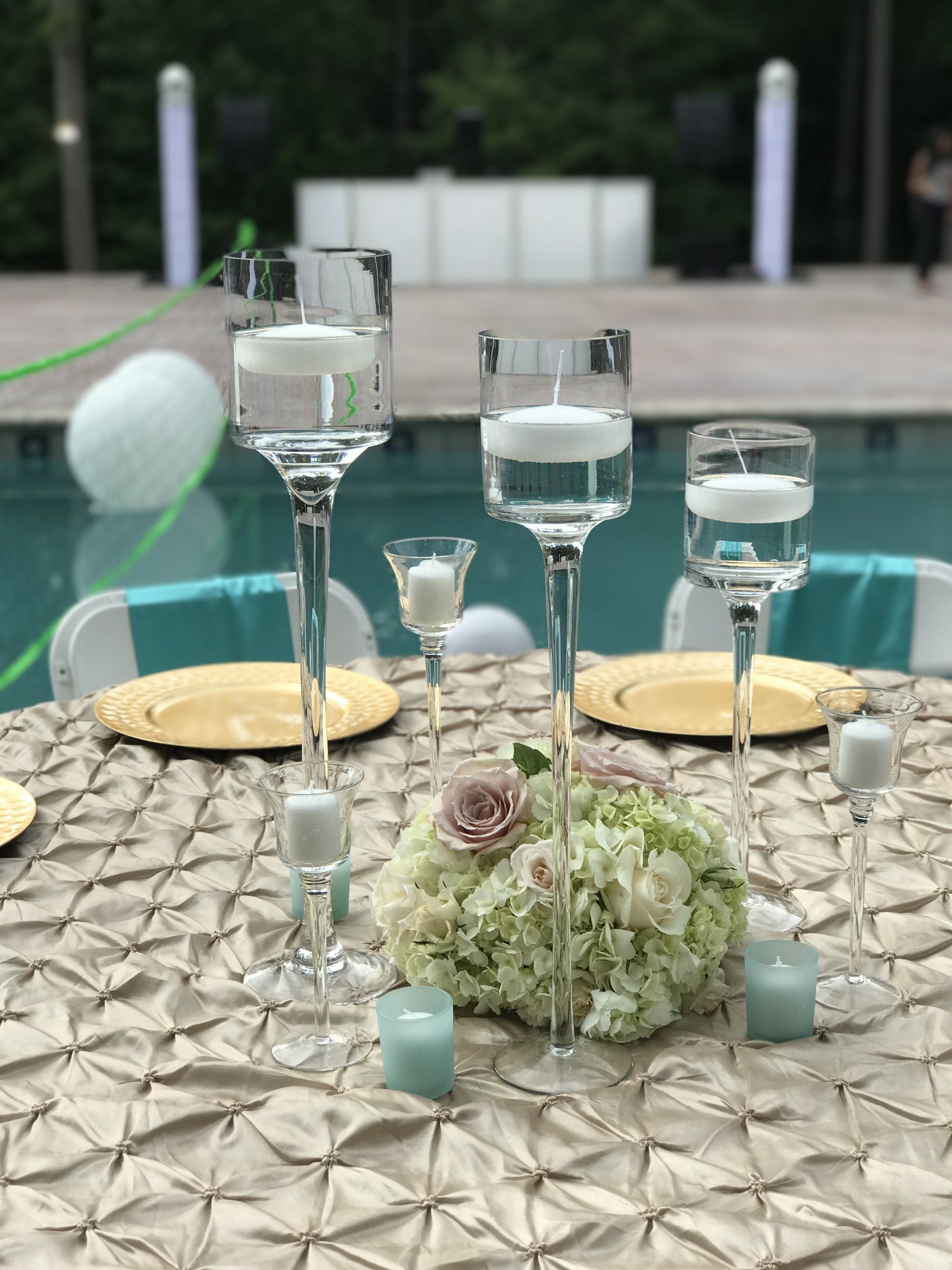 Outdoor event design with varying height tall stem floating candle holders, floral arrangement, gold chargers, and a pinwheel button table cloth.