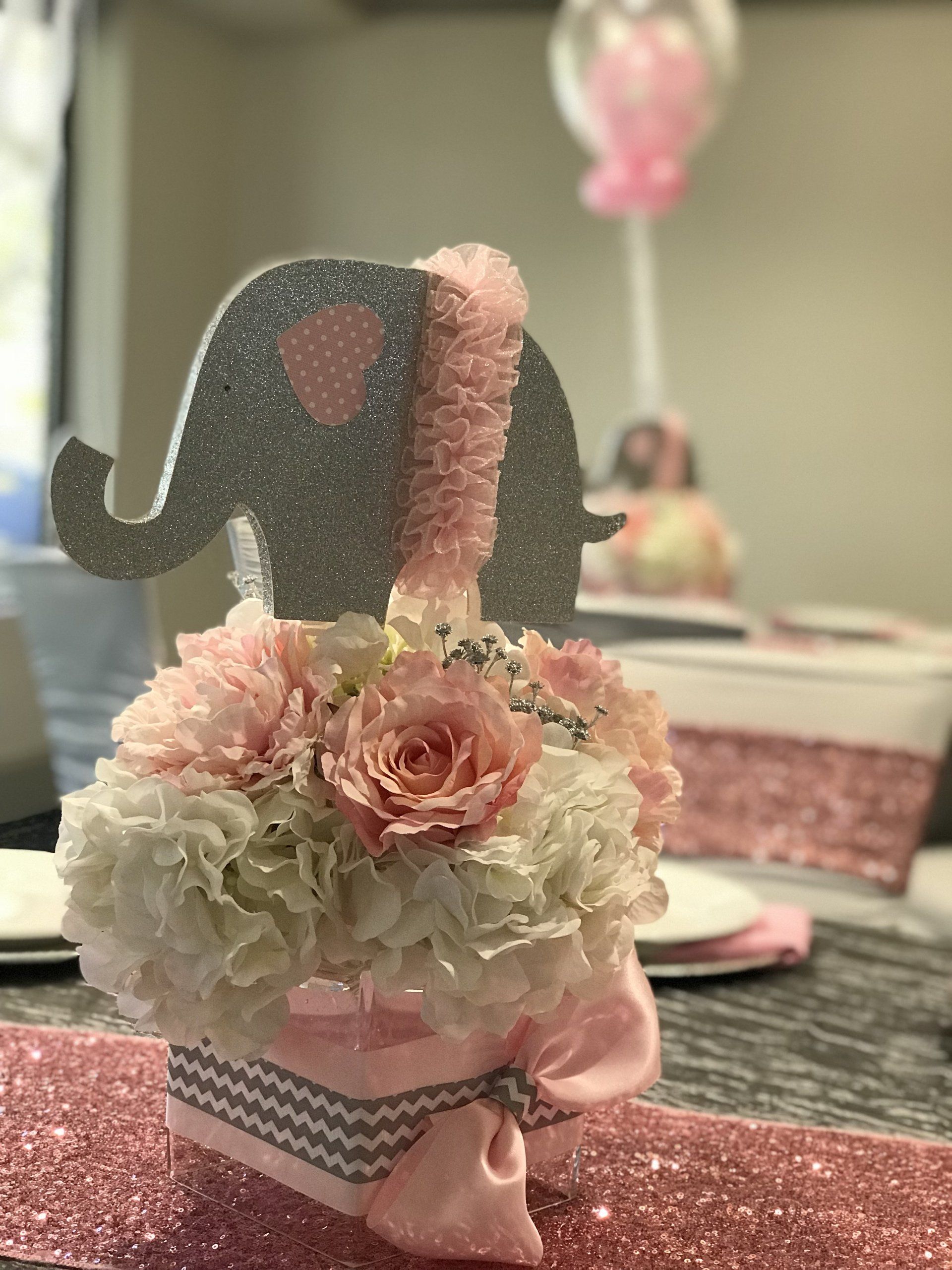 Baby shower event design with a pink, white and grey theme. Centerpiece is a floral arrangement sitting on a cube glass base and an elephant cutout on top.