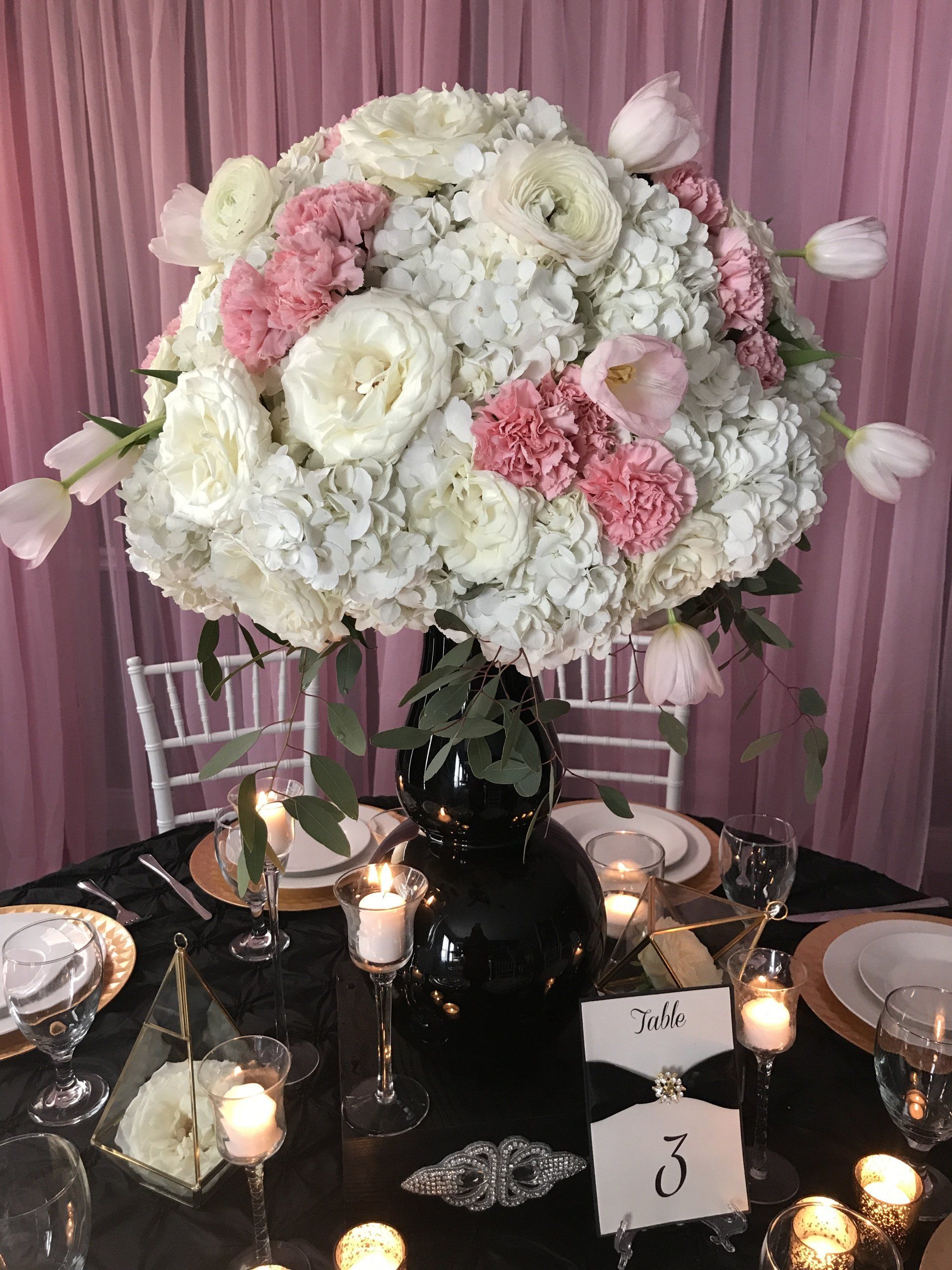 Custom centerpiece design with a tall floral arrangement sitting on an elegant box with an embellishment, gold trimmed glass prisms, a table number and various candles.