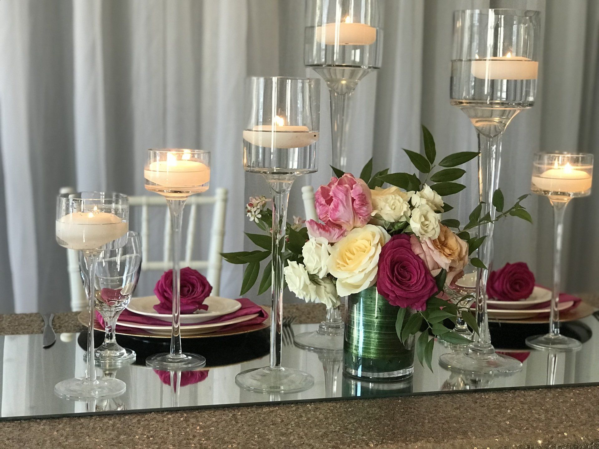 Custom designed centerpiece design with one floral arrangement in a short cylinder vase and long stem floating candle holders of varying sizes sitting on a mirror.