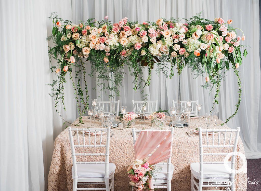 Beautiful custom event design with draping in the background, a table setup with an elevated bridge floral centerpiece, candles, and white banquet (Chiavari) chairs.