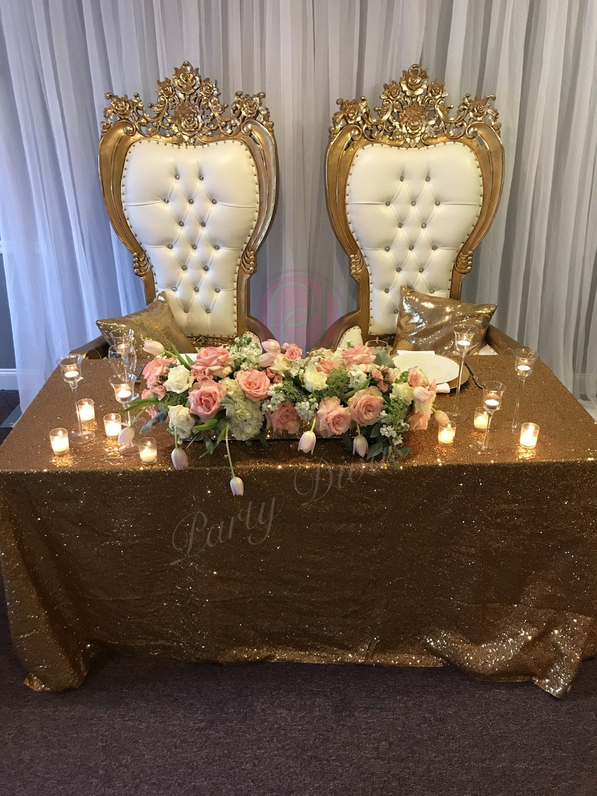 Head table custom design with various candles, a wide floral arrangement, and various candles sitting on a sparkling gold tablecloth. Gold trimmed thrones and draping are in the background.