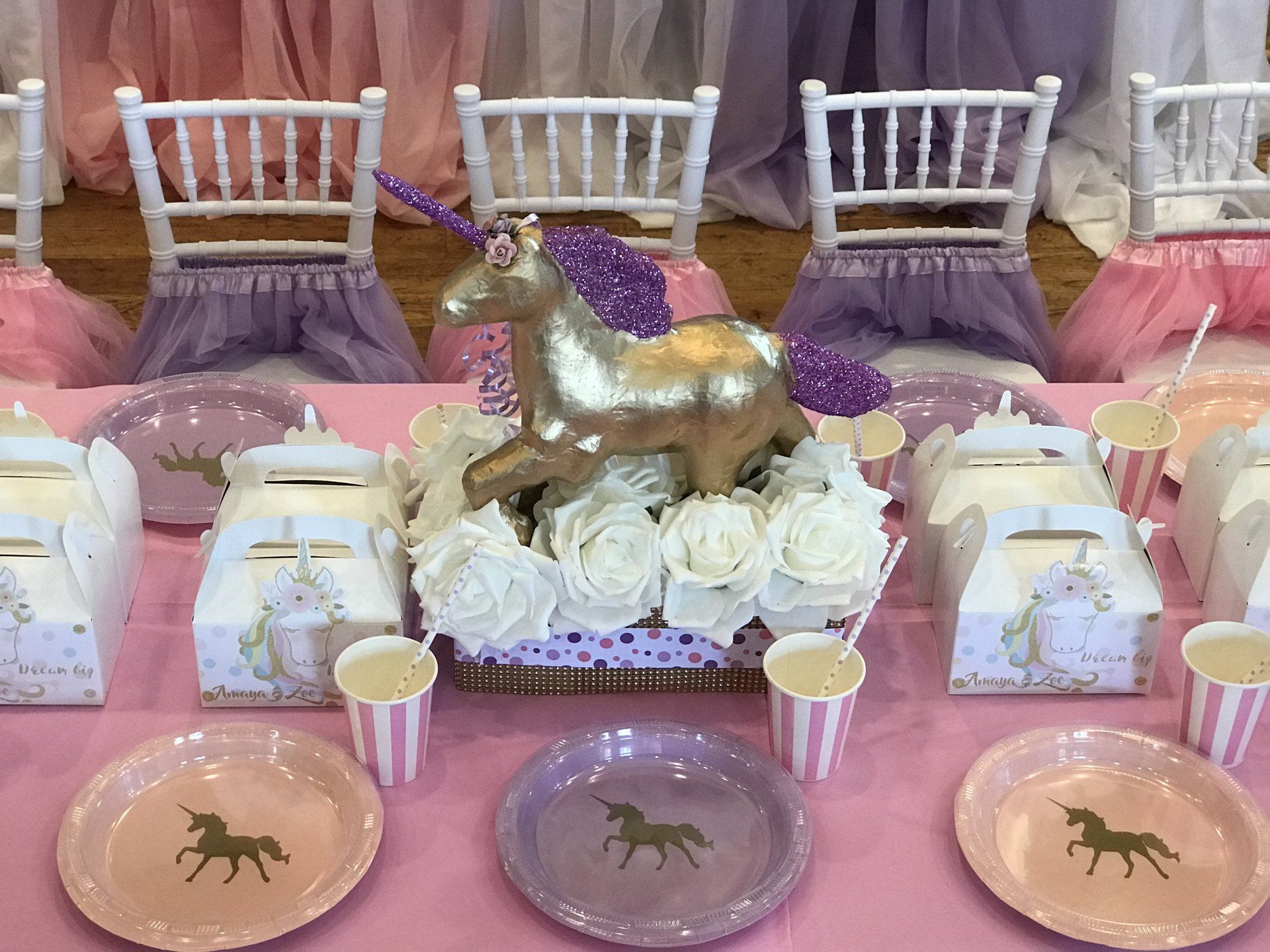 Unicorn themed table set up with a pink table cloth, fun floral centerpiece with a gold unicorn and party favors lined up in the center.